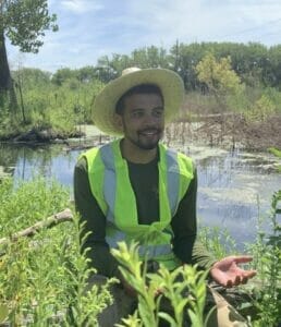 Adam Glueckert sits in front of a swampy body of water among lush native plants. Adam wears a wide-brimmed had and a green high visibility vest.